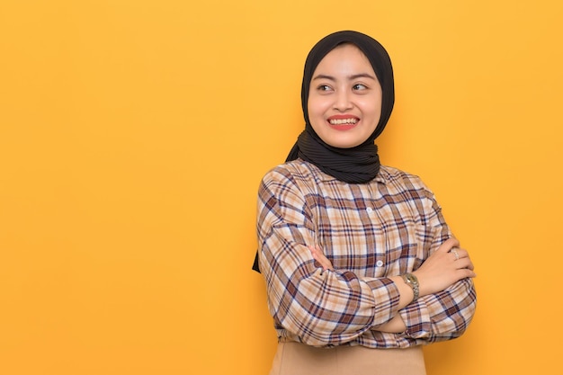 Smiling young asian woman in plaid shirt standing with crossed arms looking away at copy space isolated on yellow background