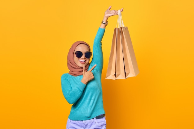 Smiling young asian woman pointing fingers at shopping bags on yellow background