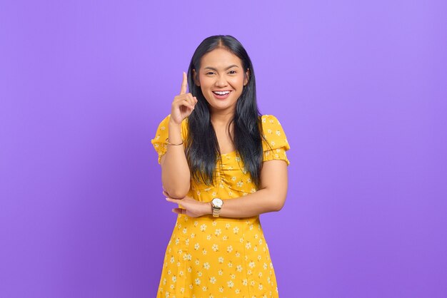 Smiling young Asian woman pointing fingers up and having an idea on purple background