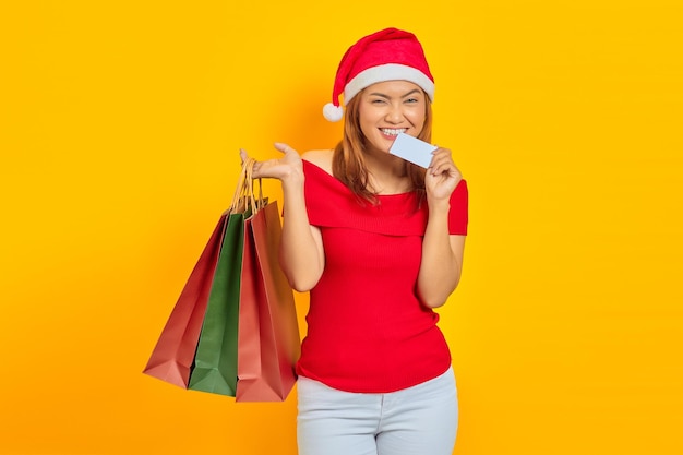 Smiling young asian woman in santa claus hat holding shopping bag and biting credit card