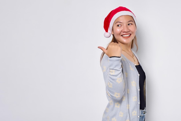 Улыбающаяся молодая азиатка в шляпе Санта-Клауса, указывающая большим пальцем на пространство для копирования, изолированное на белом фоне
