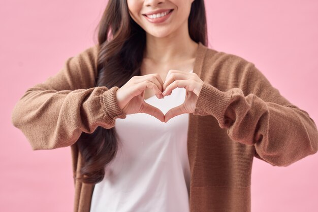 Улыбающаяся молодая азиатская женщина показывает жест сердца двумя руками и смотрит в камеру, изолированную на розовом фоне