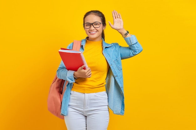 Smiling young asian woman student in denim clothes with backpack holding notebook waving and greeting with hand as notices someone isolated on yellow background