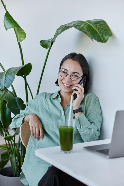 Smiling young asian woman talks via mobile phone wears round\
spectacles and shirt uses laptop for distance work drinks fresh\
green smoothie makes call poses at white desktop against potted\
plant