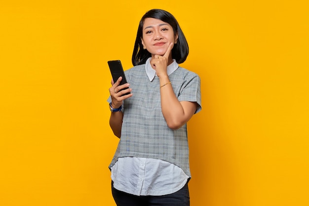 Smiling young Asian woman thinking about question with finger hand on chin and holding mobile phone