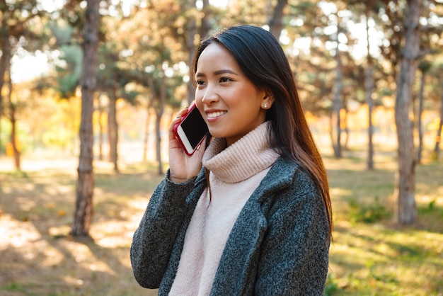 公園で屋外を歩いて、携帯電話で話しているコートを着て笑顔の若いアジアの女性