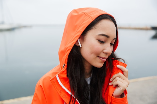 Smiling young asian woman wearing raincoat spending time outdoors walking at the coastland, listening to music with wireless earphones