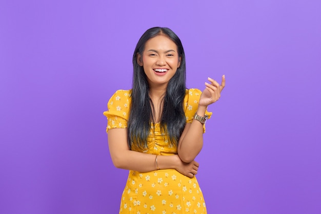 Smiling young Asian woman wearing yellow dress and looking confident on purple background