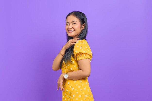 Smiling young asian woman wearing yellow dress and looking confident on purple background