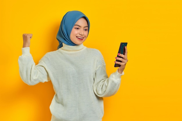 Smiling young asian woman in white sweater and hijab using
mobile phone doing winner gesture isolated over yellow background
people islam religious concept