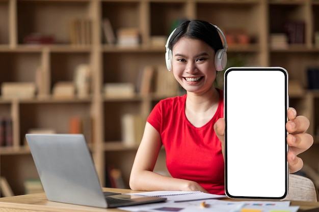 Smiling young asian woman with down syndrome in headphones with laptop shows phone with blank screen