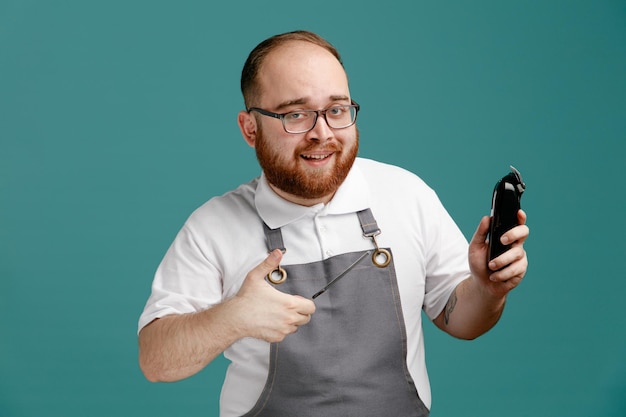 Smiling young barber wearing uniform and glasses holding teaser comb and hair trimmer looking at camera showing thumb up isolated on blue background