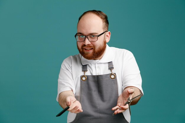 Smiling young barber wearing uniform and glasses stretching comb and scissors out towards camera while looking at camera isolated on blue background