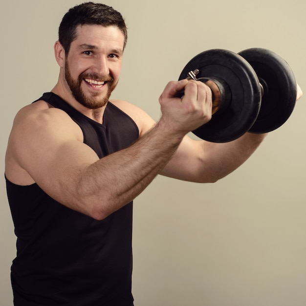 A smiling young bearded athlete in a black T-shirt raises a heavy dumbbell in front of him.