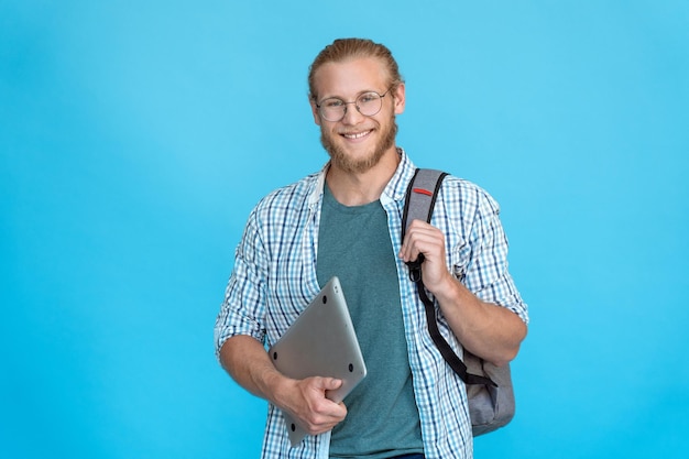 웃고 있는 젊은 수염을 기른 대학생은 안경을 쓰고 현대적인 노트북 복사 공간을 들고 있습니다.