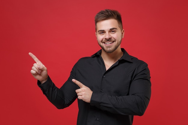 Smiling young bearded guy 20s in classic black shirt posing isolated on red background studio portrait. People sincere emotions lifestyle concept. Mock up copy space. Pointing index fingers aside up.