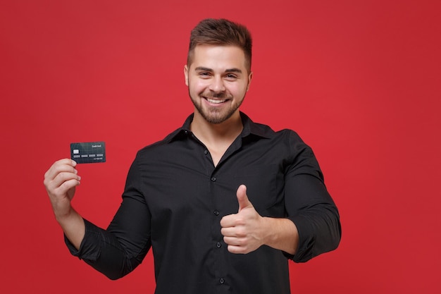 Smiling young bearded guy in classic black shirt posing isolated on bright red wall background studio portrait. People lifestyle concept. Mock up copy space. Hold credit bank card, showing thumb up.