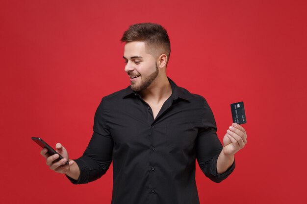 Smiling young bearded guy in classic black shirt posing isolated on red background studio portrait. People sincere emotions lifestyle concept. Mock up copy space. Hold credit bank card, mobile phone.