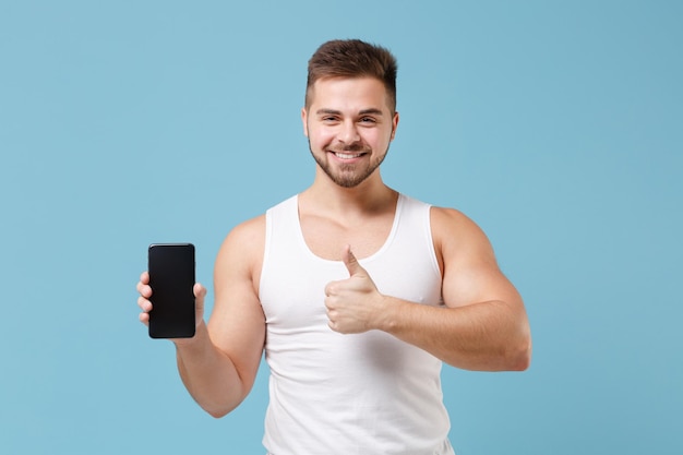 Smiling young bearded guy in white singlet isolated on pastel blue background. Sport fitness healthy lifestyle concept. Mock up copy space. Hold mobile phone with blank empty screen, showing thumb up.