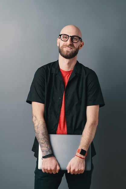 Smiling young bearded man with glasses holding laptop under his arms in the studio on gray