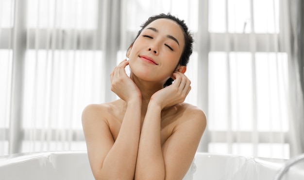 Улыбка молодой красивой азиатской женщины чистой свежей здоровой белой кожи касаясь ее лица рукой и применяя крем дома.