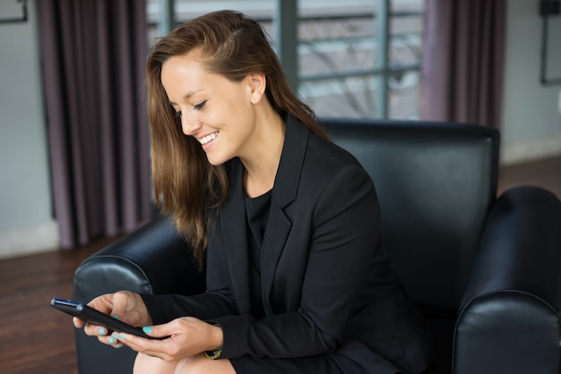 smiling young beautiful brown-haired woman using smartphone and sitting in armchair