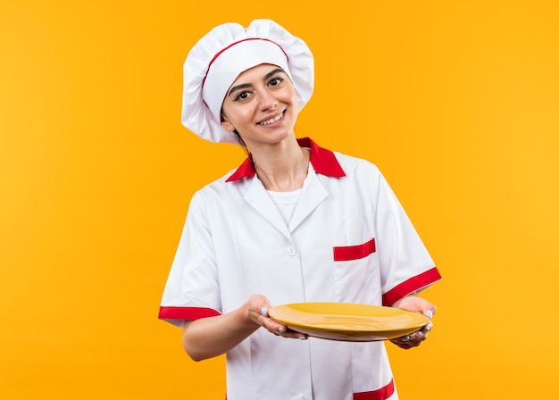 Sorridente giovane bella ragazza in uniforme da chef che porge piatto in telecamera