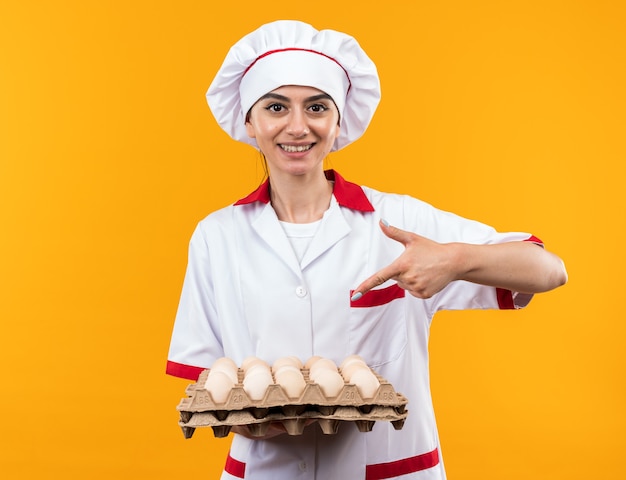 Smiling young beautiful girl in chef uniform holding and points at batch of eggs