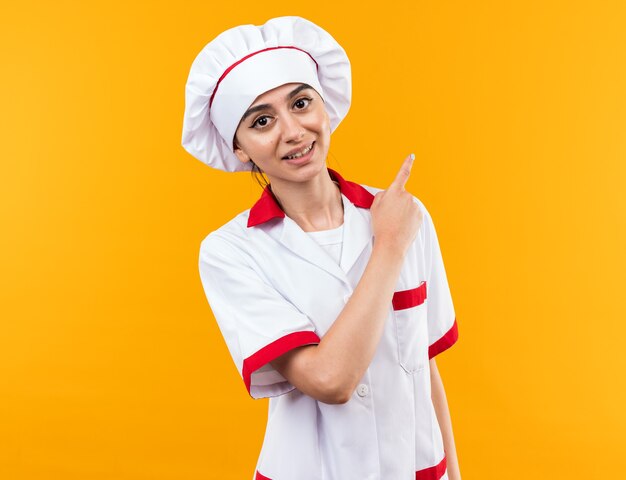 Smiling young beautiful girl in chef uniform points at behind 