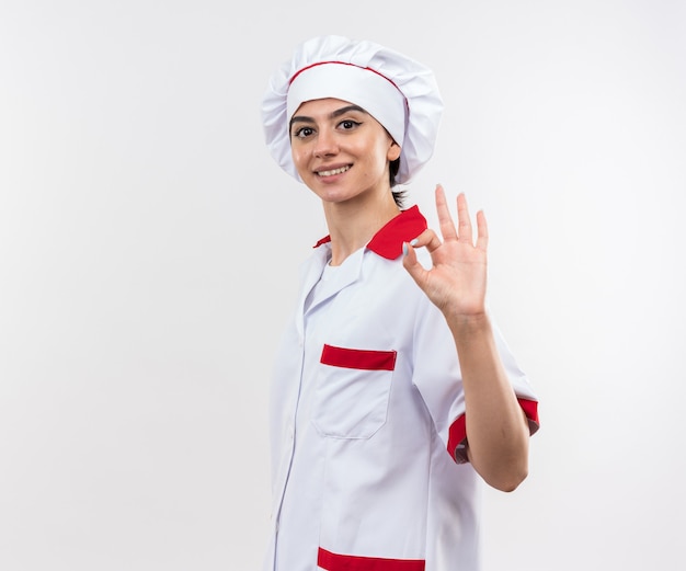 Smiling young beautiful girl in chef uniform showing okay gesture isolated on white wall with copy space