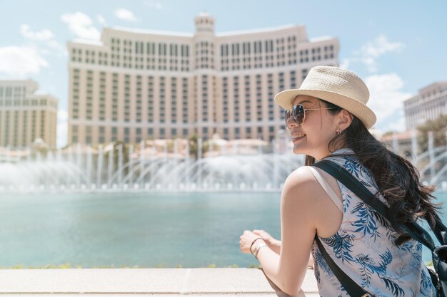 Smiling young beautiful girl traveler standing near modern\
fountain with tall hotel building in background. woman tourist\
watching sights of seeing outdoor on sunny day in summer las vegas\
usa.