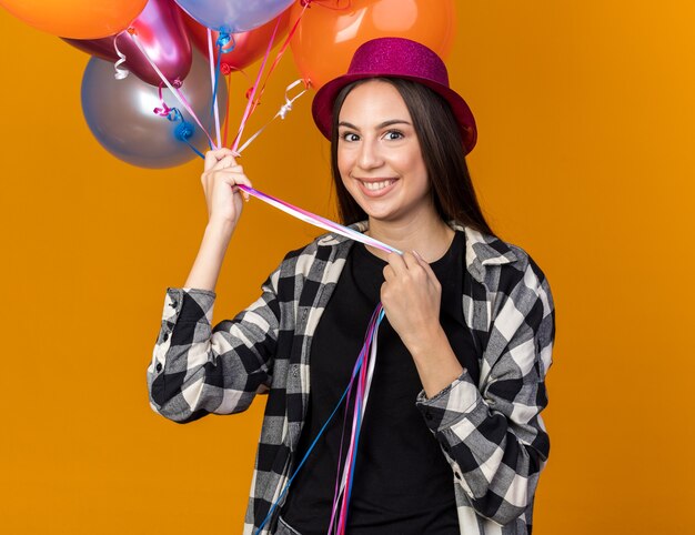 Smiling young beautiful girl wearing party hat holding balloons isolated on orange wall