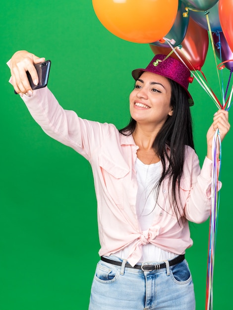 La giovane bella ragazza sorridente che indossa il cappello del partito che tiene i palloni prende un selfie