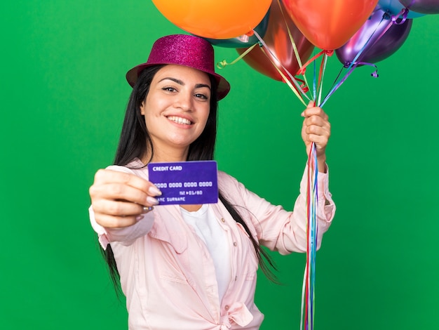 Smiling young beautiful girl wearing party hat holding balloons with credit card isolated on green wall