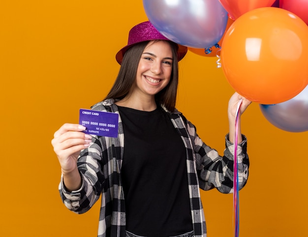 Smiling young beautiful girl wearing party hat holding balloons with credit card isolated on orange wall