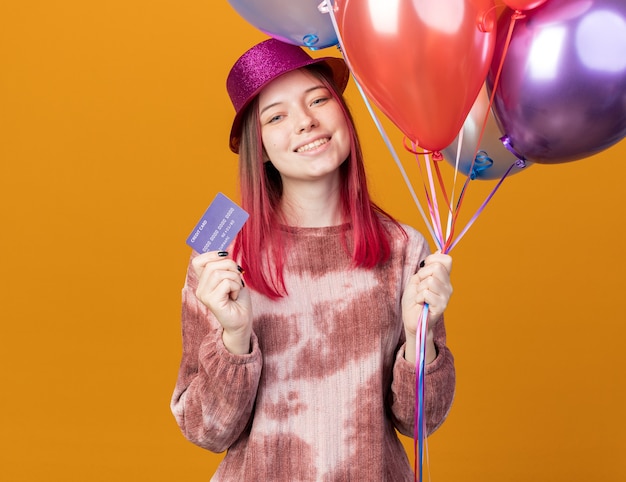 Photo smiling young beautiful girl wearing party hat holding balloons with credit card