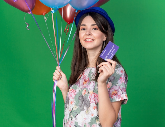 Smiling young beautiful girl wearing party hat holding balloons with credit card 