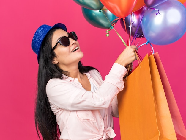 Smiling young beautiful girl wearing party hat holding balloons with gift bags isolated on pink wall