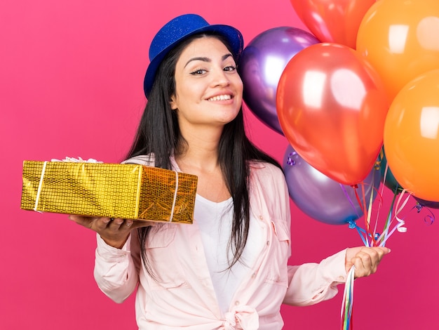 Sorridente giovane bella ragazza che indossa un cappello da festa con palloncini con scatola regalo