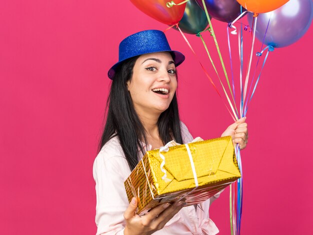 Smiling young beautiful girl wearing party hat holding balloons with gift box 