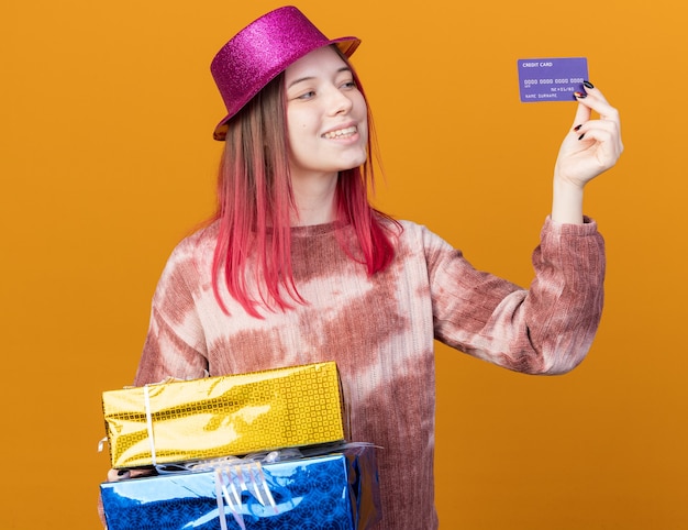 Smiling young beautiful girl wearing party hat holding gift boxes and looking at credit card in her hand 