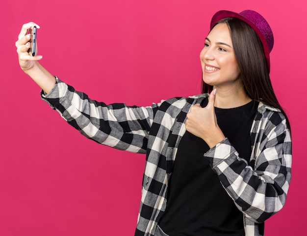 Smiling young beautiful girl wearing party hat take a selfie showing thumb up isolated on pink wall