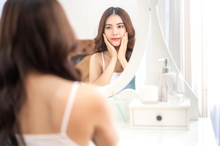 若い美しいきれいなアジアの女性の笑顔は、手で彼女の顔に触れて、homespaと美しさの概念でクリームを適用するミラーアジアの女の子を見てきれいな新鮮な健康的な白い肌