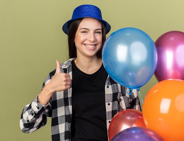 Smiling young beautiful woman wearing blue hat standing nearby balloons showing thumb up isolated on olive green wall