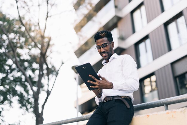 通りで彼のタブレットに取り組んでいる白いシャツを着た若い黒人ビジネスマンの笑顔