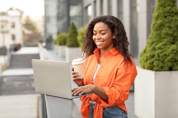 Smiling young black lady with cup of coffee to go chatting on\
computer working remotely has video call