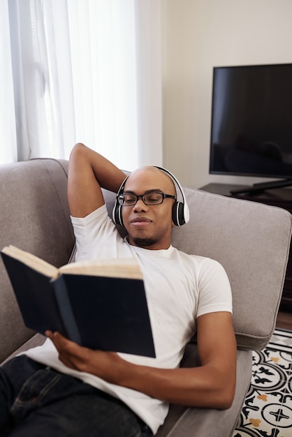 Smiling young Black man in headphones relaxing on sofa and reading interesting novel