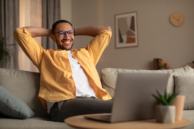 Photo smiling young black man with laptop relaxing after working online sitting on couch with laptop at