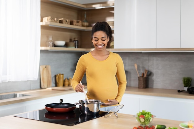 Smiling young black pregnant female with big belly preparing\
dinner and salad in minimalist kitchen interior