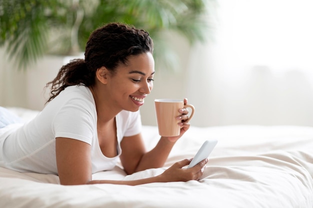 Smiling young black woman resting in bed with smartphone and coffee